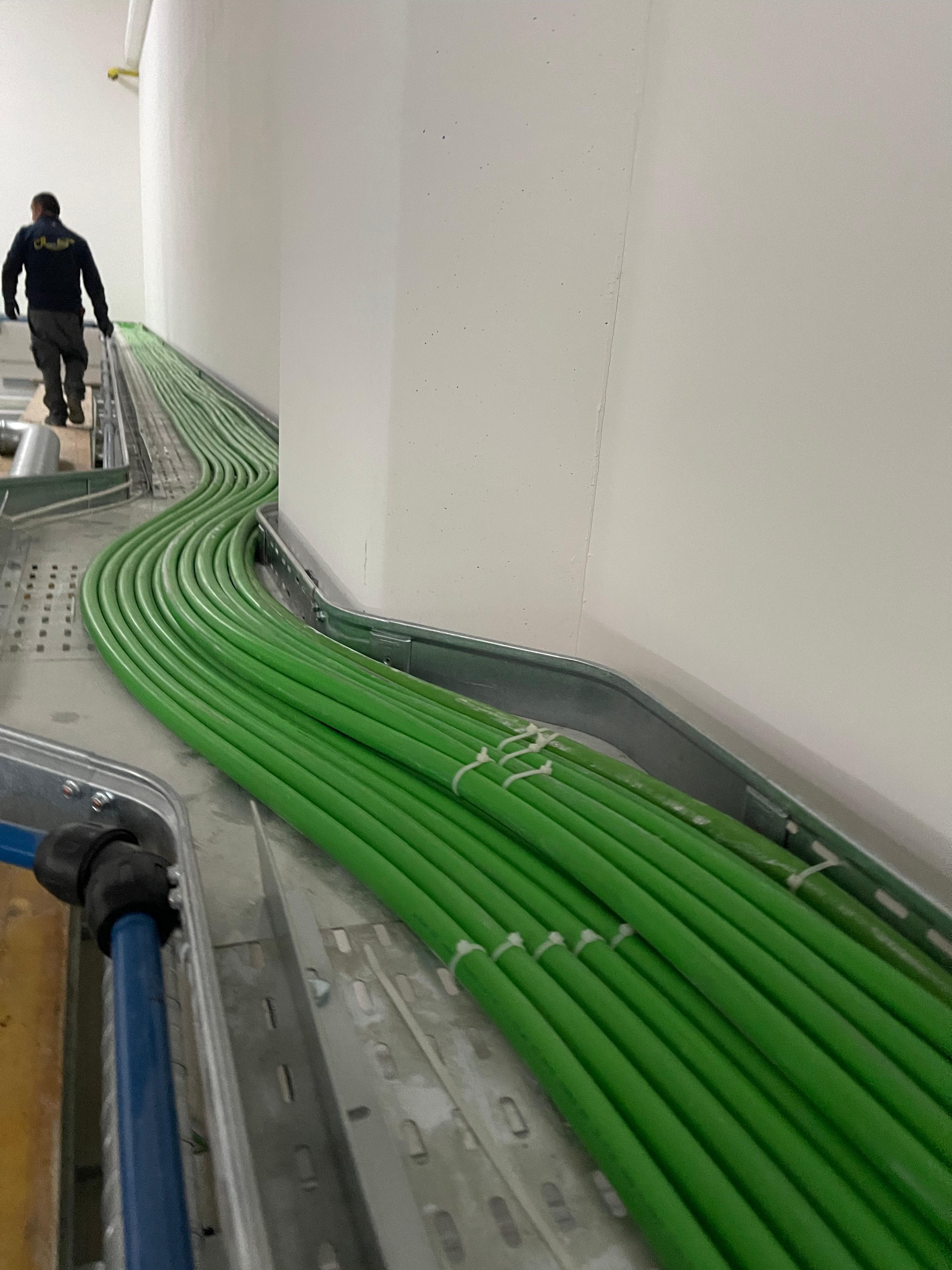 Cavi verdi disposti ordinatamente in una canalina metallica curva lungo una parete bianca industriale.