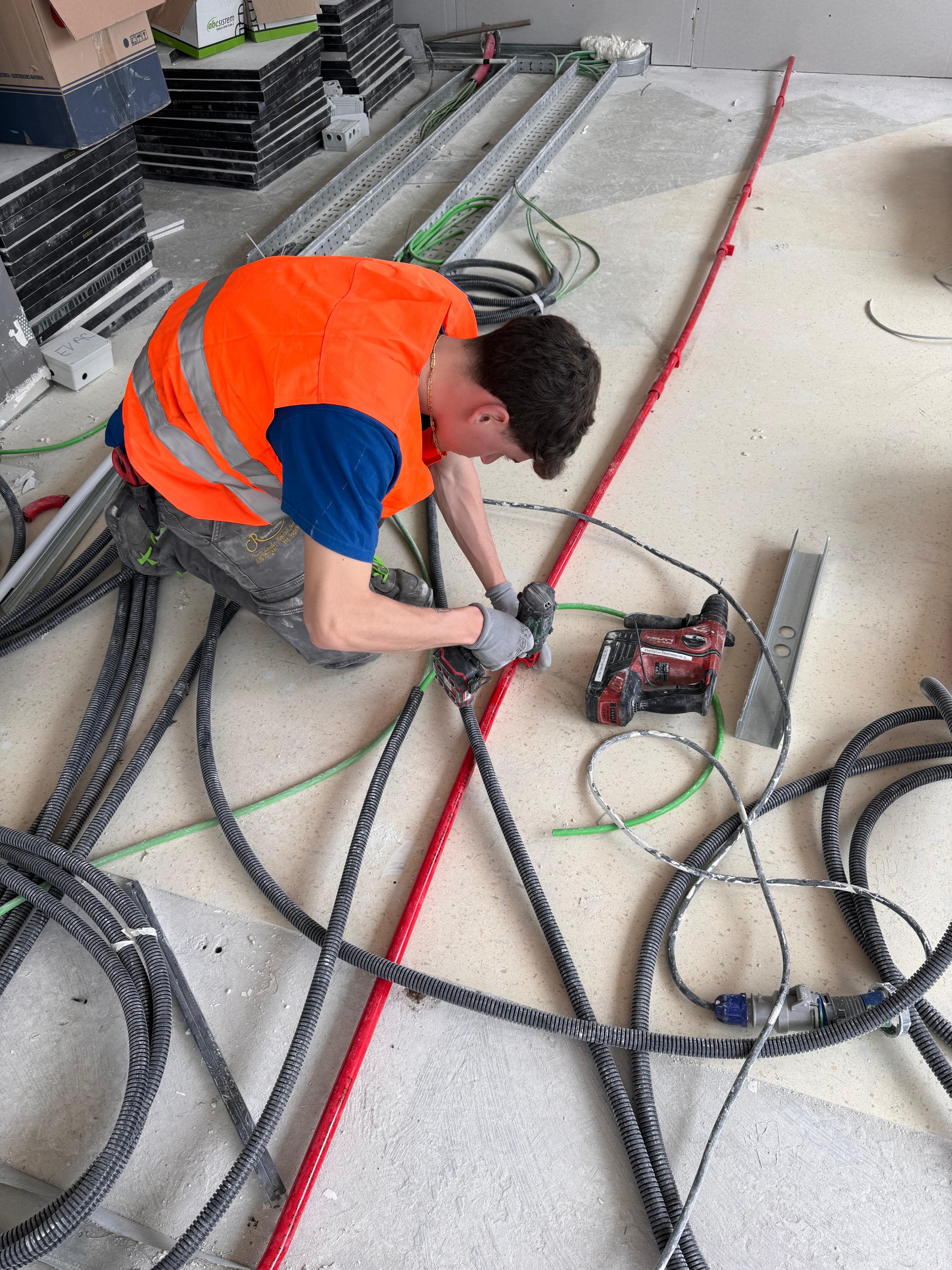 Operaio edile in gilet arancione che lavora con un trapano tra cavi e tubazioni.
