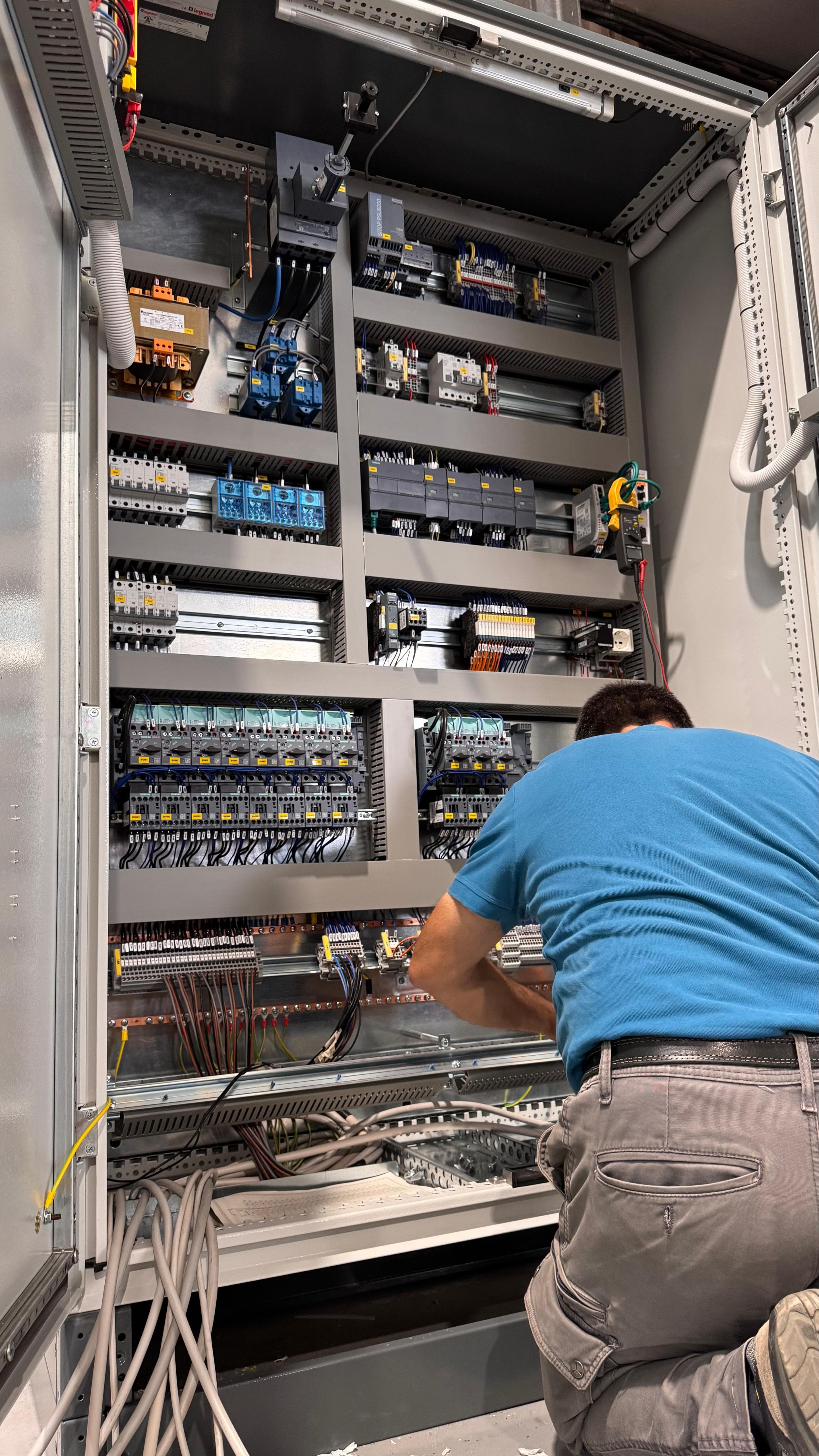 Tecnico in maglietta blu che lavora su un complesso quadro elettrico industriale con molti cablaggi.